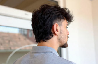 Homme de profil avec un mulet moderne, cheveux noirs ondulés, côtés courts, arrière long, barbe et boucle d'oreille.